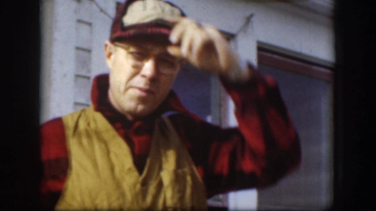 Middle-aged man in red-and-black plaid flannel, yellow canvas vest, and thick-framed glasses adjusts his two-tone work cap while speaking to camera; exterior clapboard house, Kansas, circa 1958, 8mm.