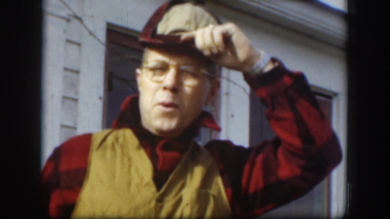 Same man tips his two-tone cap with right hand, mouth open mid-speech; clapboard house siding and window visible behind him.