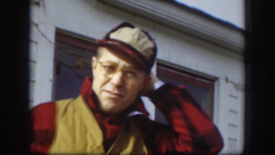 Man in red plaid flannel shirt and yellow canvas work vest pauses mid-gesture with cap repositioned; exterior clapboard house, Kansas, circa 1958, 8mm home movie.