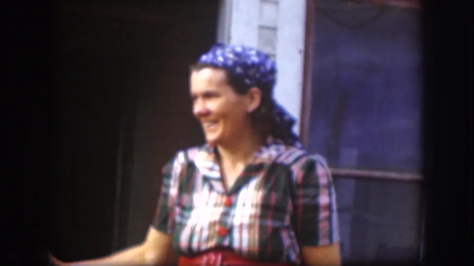 Smiling woman in late-1950s dress — plaid shirt and blue headscarf — standing near a building exterior, likely the United States circa 1958.