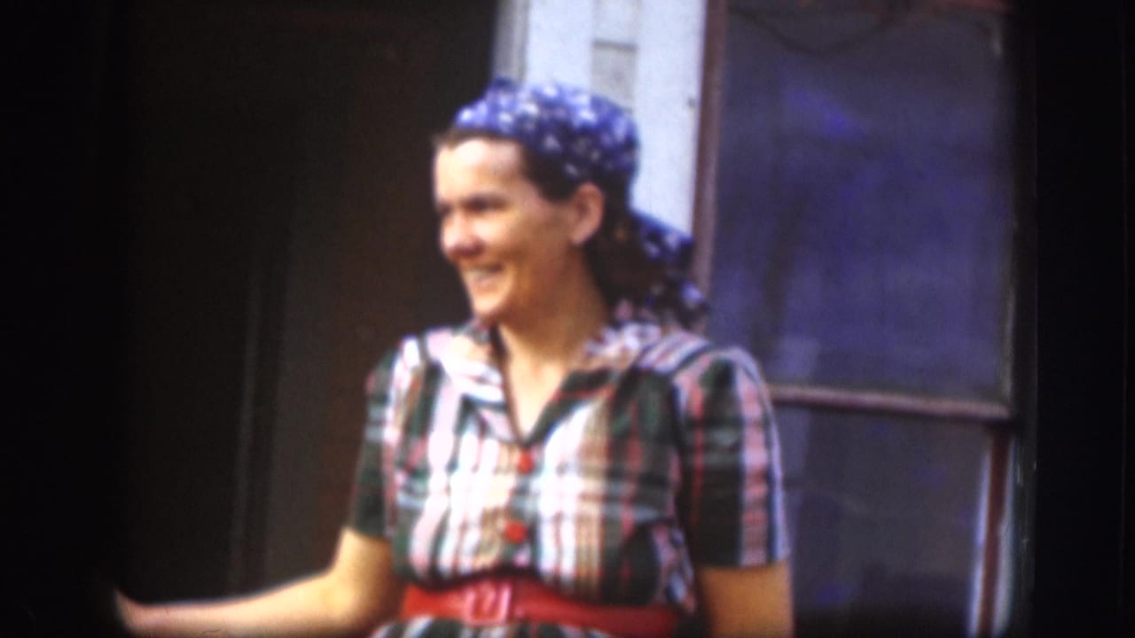 Smiling woman in a plaid short-sleeve dress, blue printed bandanna, and red belt, photographed waist-up against a blue painted surface — Kansas, 1958, 8mm home movie