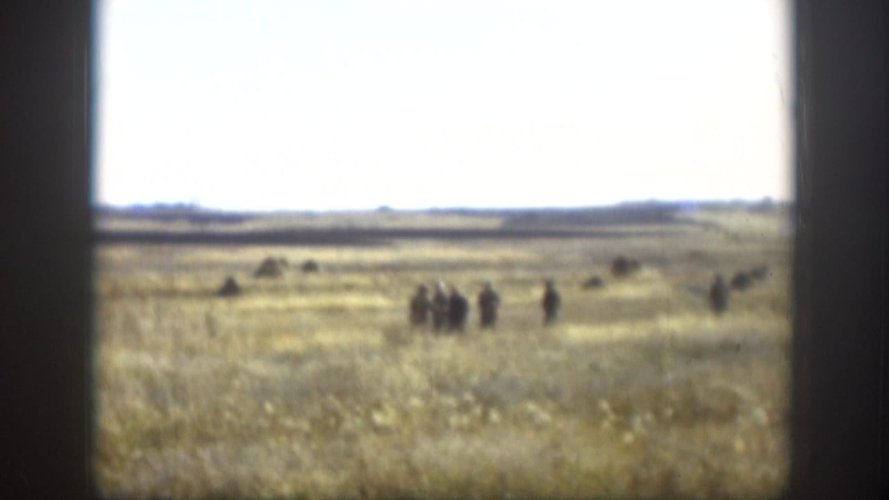 Open Kansas prairie with dry golden grass, group of people walking in the middle distance, flat horizon, and overcast pale sky — likely 1958, 8mm home movie