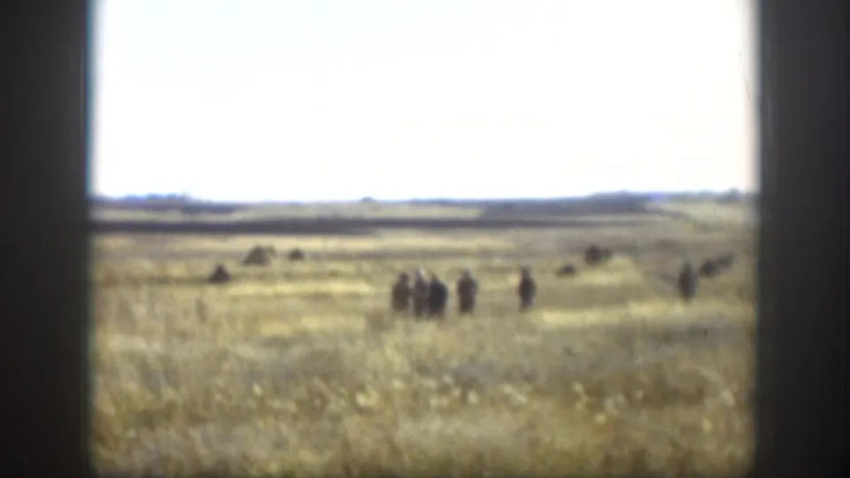 Open Kansas prairie with dry golden grass, group of people walking in the middle distance, flat horizon, and overcast pale sky — likely 1958, 8mm home movie