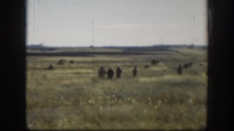 Wide Kansas grassland with scattered hay mounds, small group of figures walking in mid-distance, partial color desaturation and film scratches — likely Kansas, 1958, 8mm
