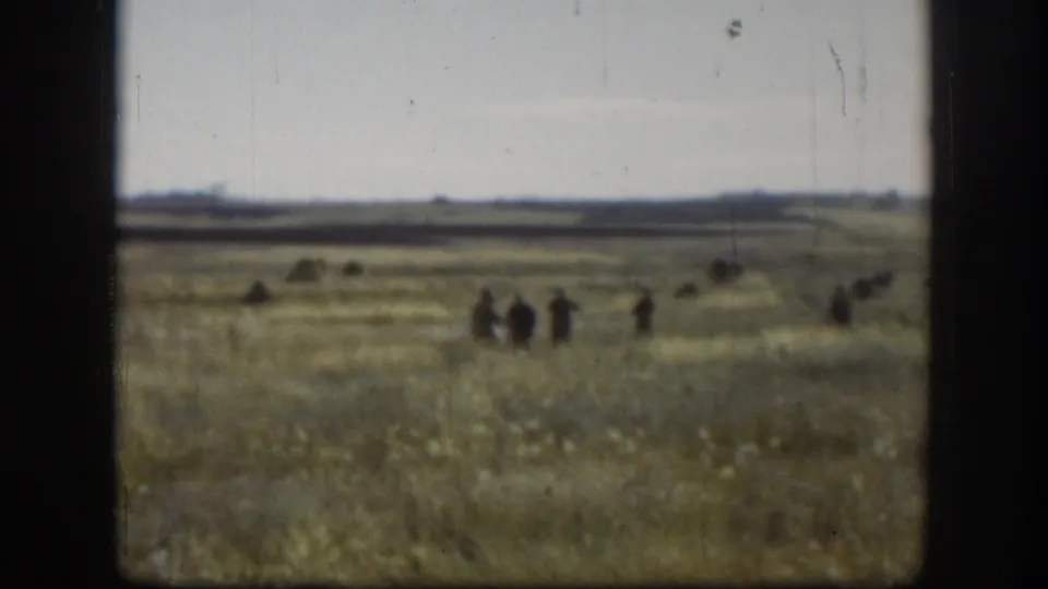 Flat Kansas prairie with dry grass, scattered hay mounds, small group of people walking in mid-distance, heavy film grain and overcast sky — likely 1958, 8mm home movie