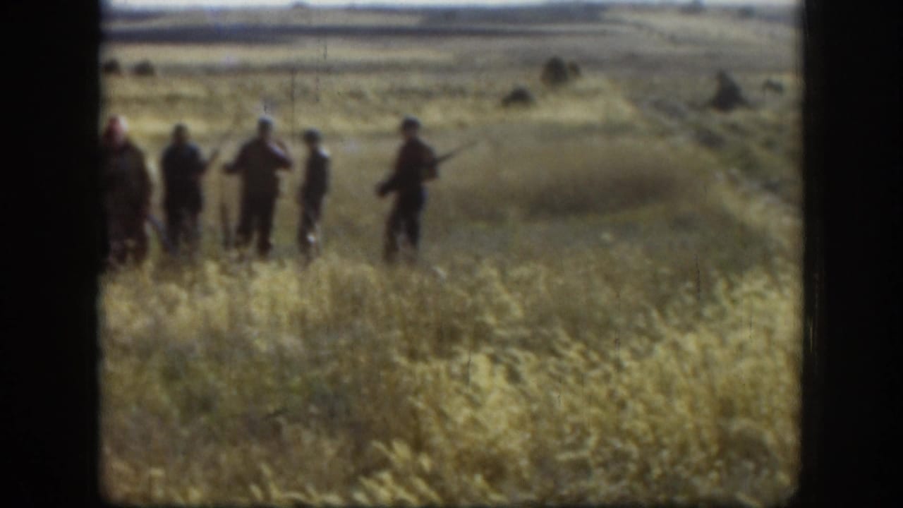 Group of men standing in tall golden grass, several holding long firearms, shot from closer range.