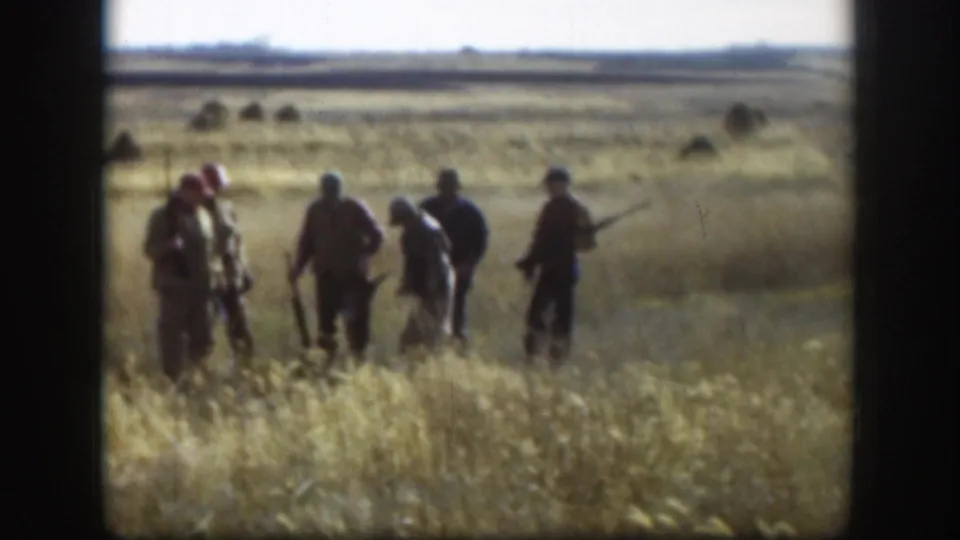 Five or six hunters grouped in dry Kansas prairie grass, c.1958; one wears a red cap; flat horizon and dark shrub clumps visible in the distance.
