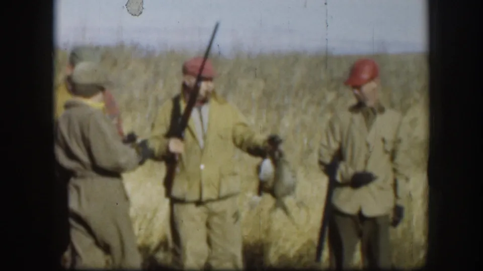 Close shot of three hunters in olive jackets and red caps handling harvested game birds in a Kansas grass field, circa 1958.