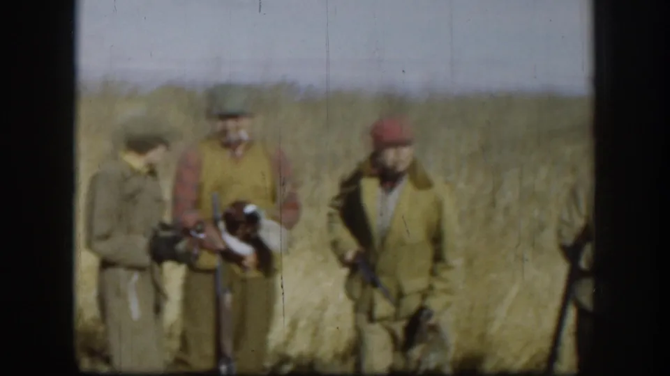 Three hunters in tan and olive jackets standing near hay bales in a Kansas field, circa 1958; vertical film scratches visible across the frame.