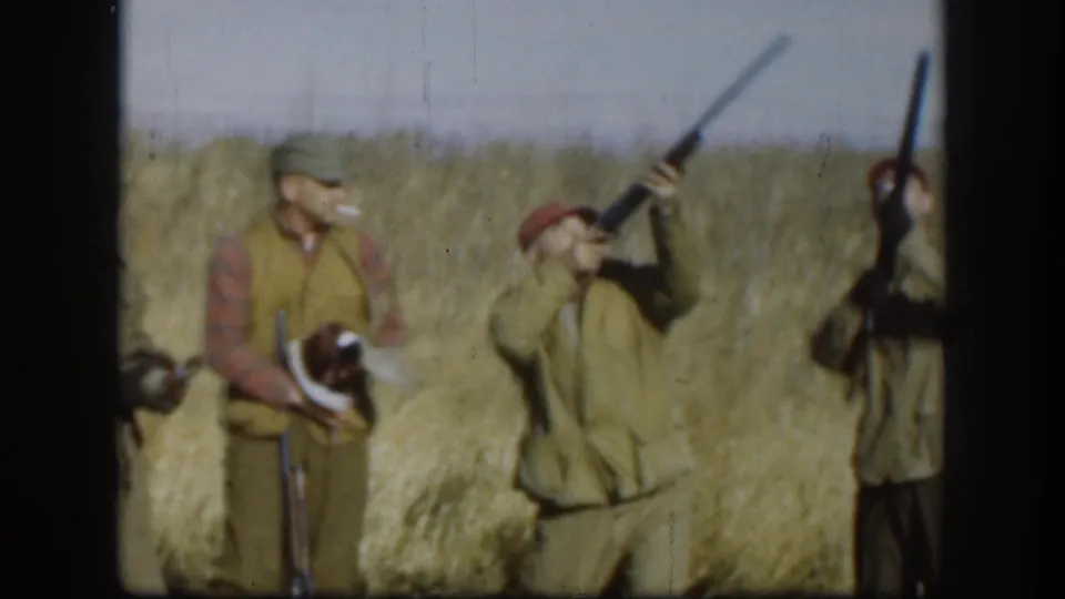 Hunters in a Kansas field circa 1958; one man appears to be smoking, another holds a shotgun raised upright, and a harvested bird is visible nearby.