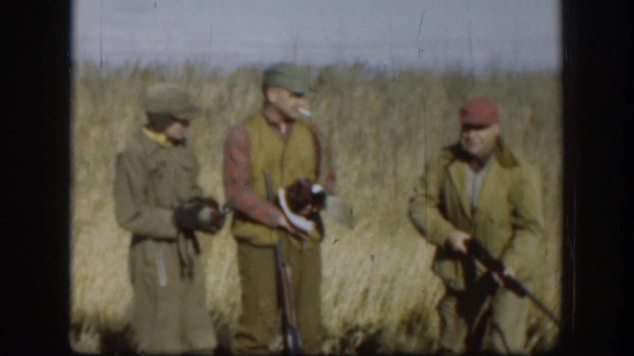 Three men in olive jackets and caps stand in a golden Kansas grassfield circa 1958, two holding shotguns at rest.