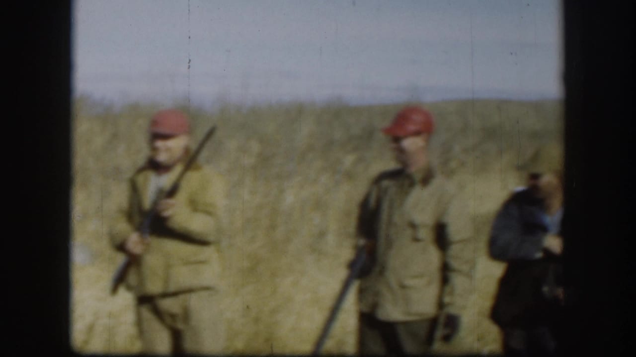 Two men in red berets and olive jackets standing in a field, one holding a shotgun upright.