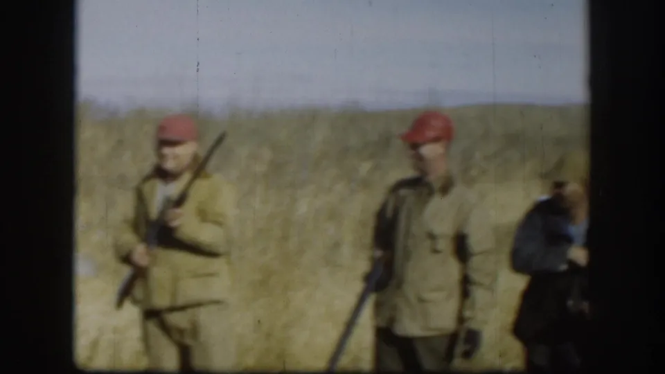 Two men in red berets and olive jackets stand in a golden grass field circa 1958 Kansas, one holding a shotgun upright.