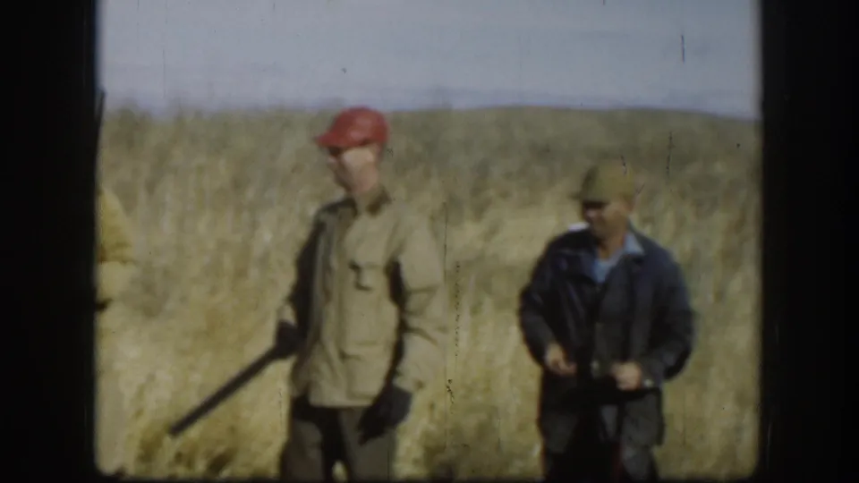 Two men in a golden grass field circa 1958, one in a red beret with a shotgun, one in a dark jacket and sunglasses; low hills visible on horizon.