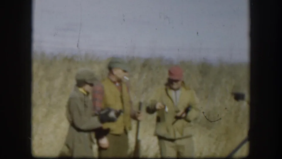 Three men in olive and plaid clothing handle shotguns in a dry grassfield circa 1958; one smokes a cigarette; film dust marks are visible.