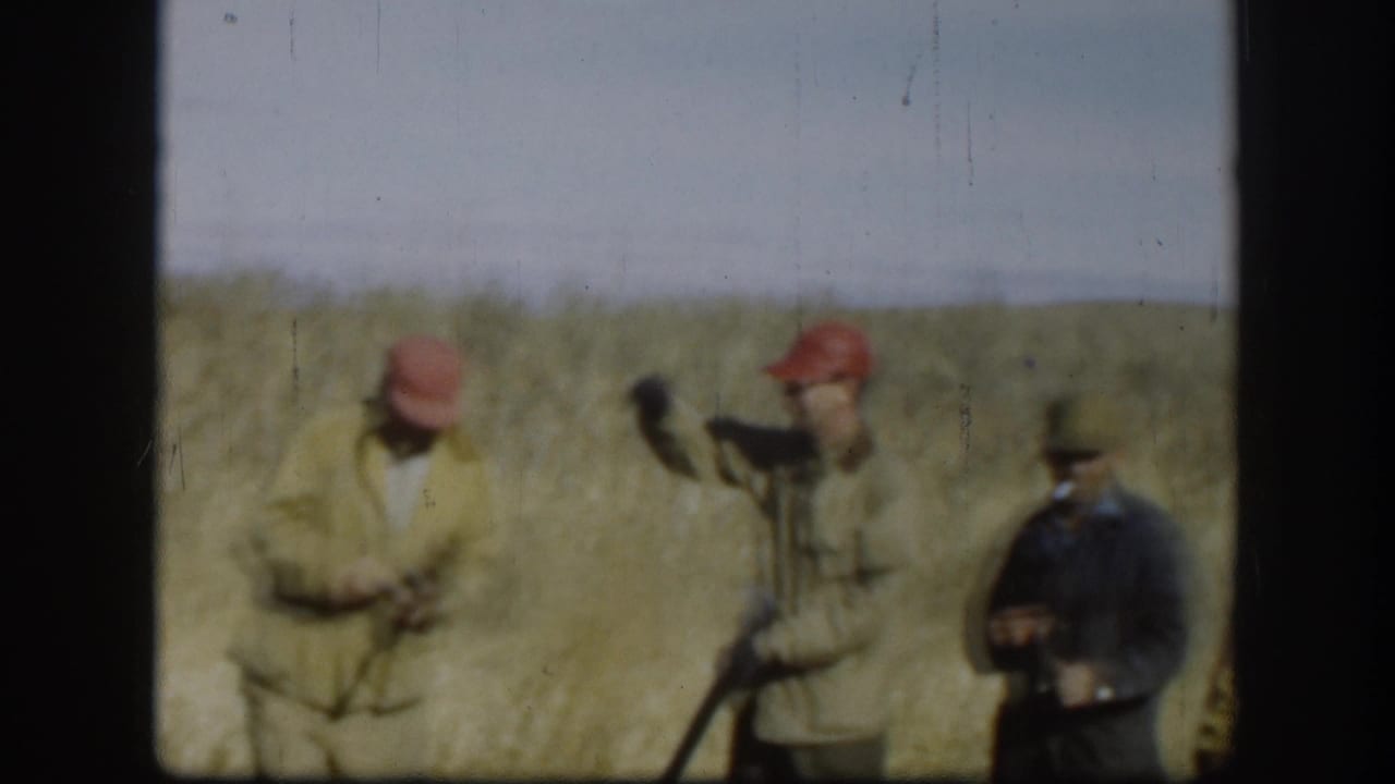 Three men in blaze-orange caps and field coats gathered in a Kansas prairie field, circa 1958; one appears to handle a shotgun.