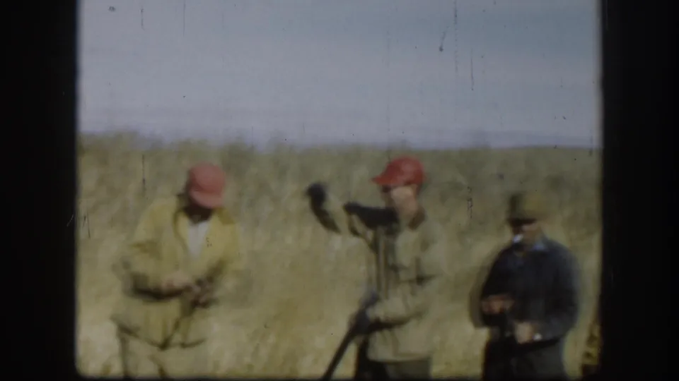 Three men in blaze-orange caps and field coats gathered in a Kansas prairie field, circa 1958; one appears to handle a shotgun.