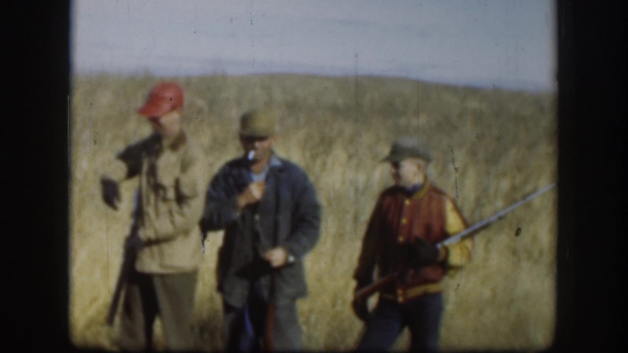 Three men in red caps and field coats conversing in a Kansas prairie field, circa 1958; a shotgun is clearly held at the right of frame.