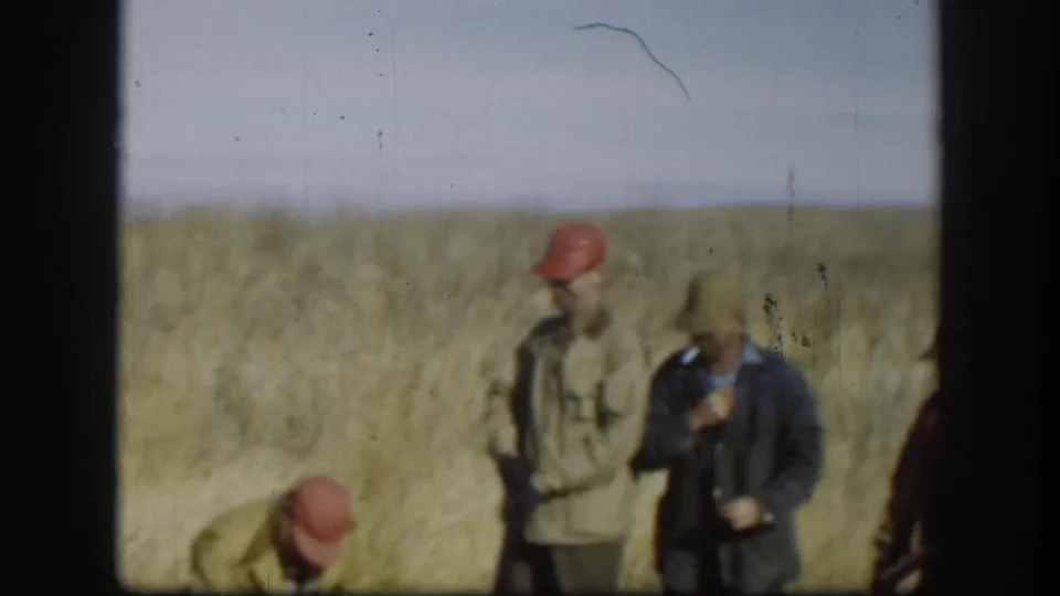 Two men in red caps in a Kansas prairie field, circa 1958; one crouches while another handles what may be a firearm; a film scratch artifact bisects the upper portion of the frame.