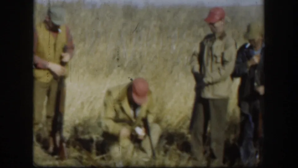 Group of hunters in blaze-orange caps amid tall Kansas prairie grass, circa 1958; one figure crouches close to the ground while another stands with a long gun.
