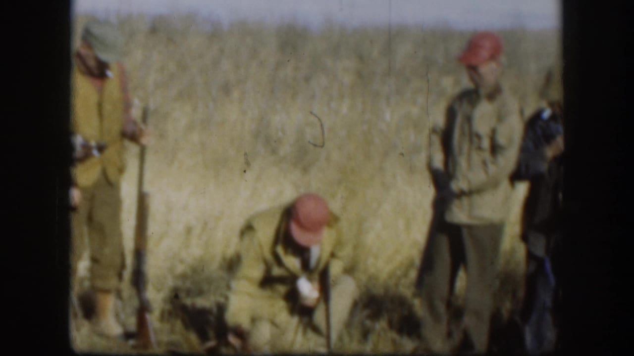 Two hunters in red-orange caps and tan jackets stand in a golden dry-grass field; one crouches while the other stands holding a rifle.