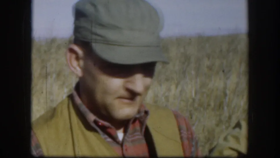 Hunter in olive cap, plaid shirt, and hunting vest with head tilted slightly down, golden Kansas grassland behind him, circa 1958.