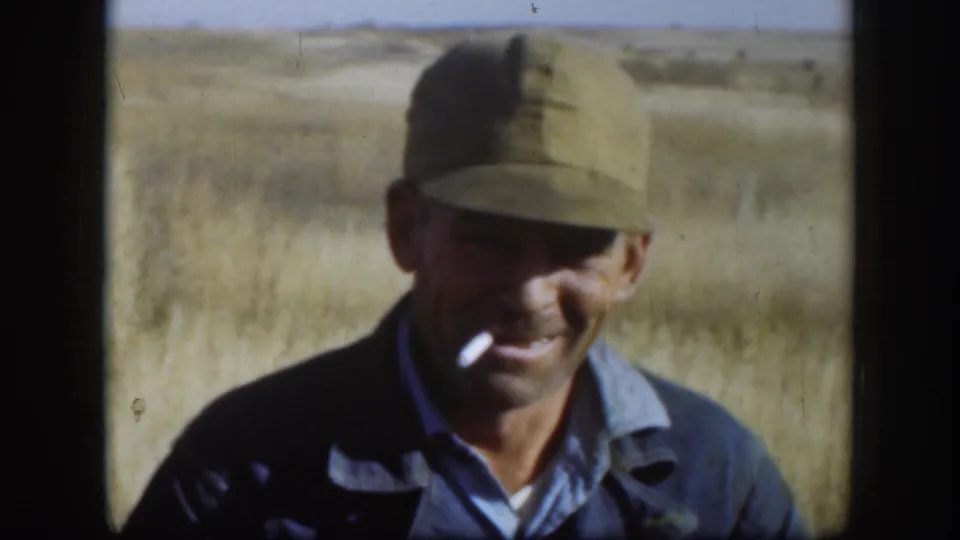 Hunter with cigarette in mouth, smiling in an open Kansas field with flat horizon and pale sky, circa 1958; darker jacket visible in this shot.
