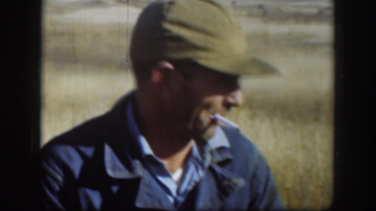 Man in khaki flat cap and dark denim jacket, seen in close profile, cigarette in mouth, against dry golden grassland.
