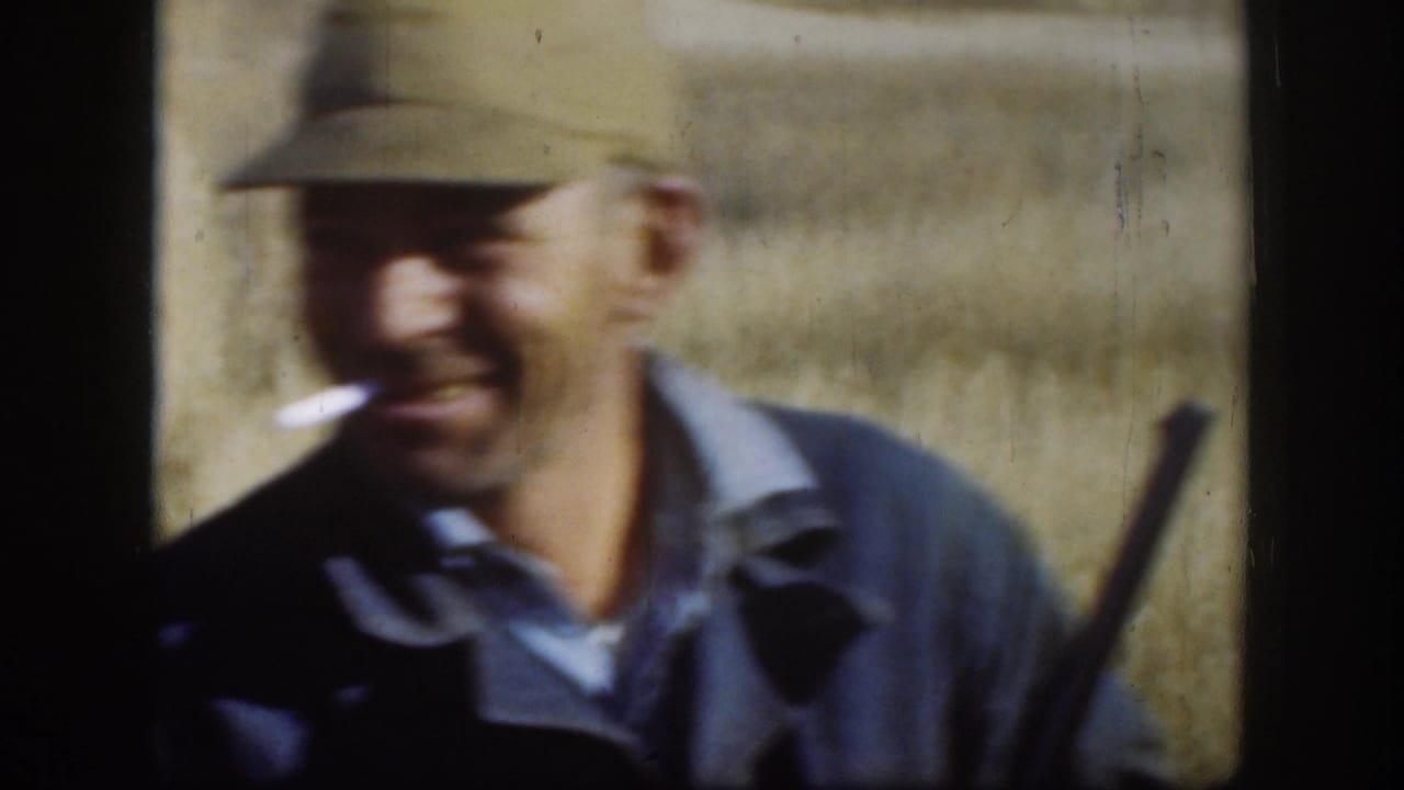 Man in khaki flat cap and dark denim jacket laughing toward camera with cigarette, rifle barrel at frame edge, in a Kansas field circa 1958 on 8mm color film.