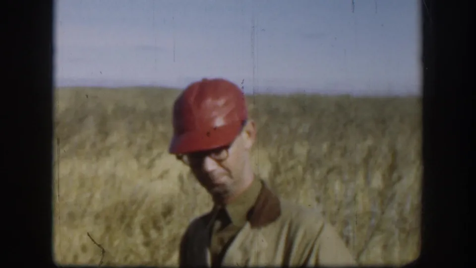 Man in red hunting cap and tan jacket looking downward in flat dry grassland with open grey-blue sky above, Kansas circa 1958, on 8mm color film.