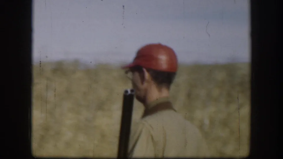 Rear three-quarter view of a man in a red hunting cap carrying a rifle upright through flat golden grassland under clear sky, Kansas circa 1958, on 8mm color film.