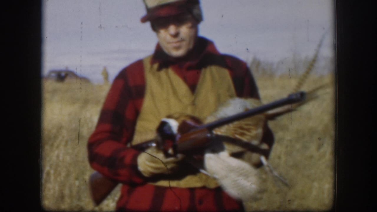 Hunter facing camera in red plaid shirt and tan vest, holding rifle and pheasant in dry grass field.