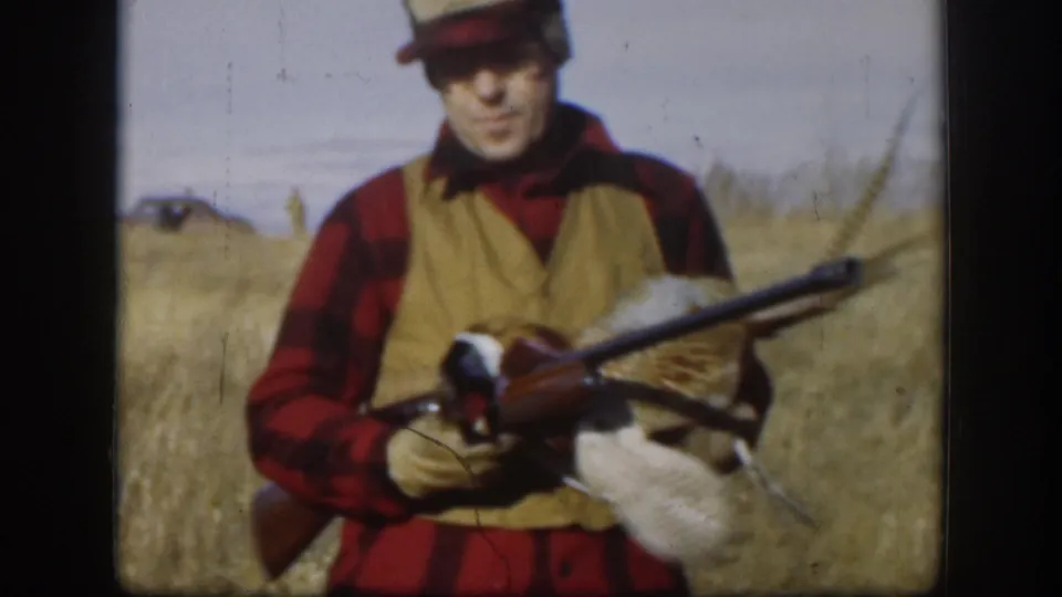 Adult male hunter displaying harvested pheasant and rifle, wearing red plaid shirt and tan hunting vest, in open Kansas grassland, circa 1958.
