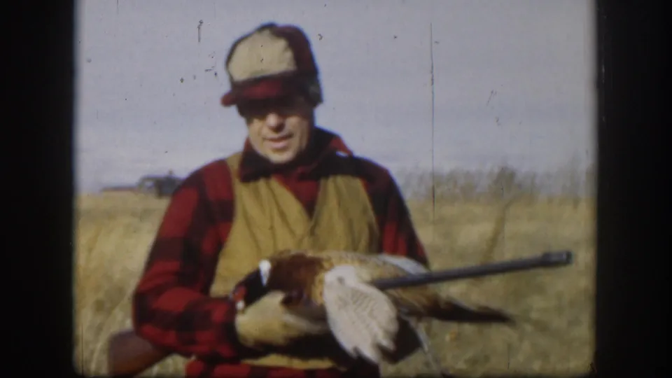 Smiling adult male hunter in red plaid shirt and tan vest holding a harvested pheasant close to camera in a Kansas prairie field, circa 1958.