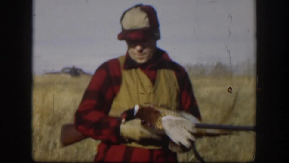 Adult male hunter in red plaid shirt and tan vest displaying a harvested pheasant in open Kansas prairie, circa 1958, with flat horizon and pale sky behind.