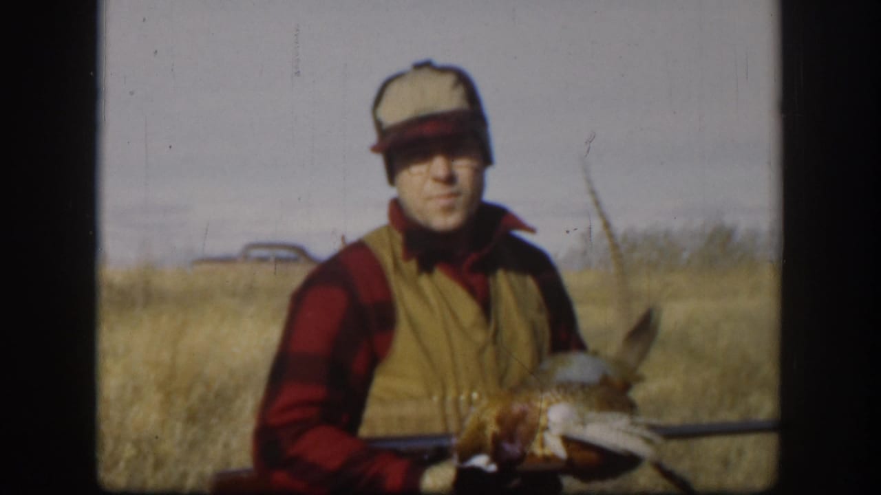 Man in 1950s hunting attire — red and black plaid shirt, tan vest, light cap — holds a harvested pheasant in a dry Kansas prairie field, circa 1958.