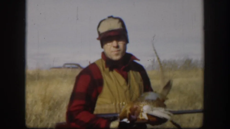 Man in 1950s hunting attire — red and black plaid shirt, tan vest, light cap — holds a harvested pheasant in a dry Kansas prairie field, circa 1958.
