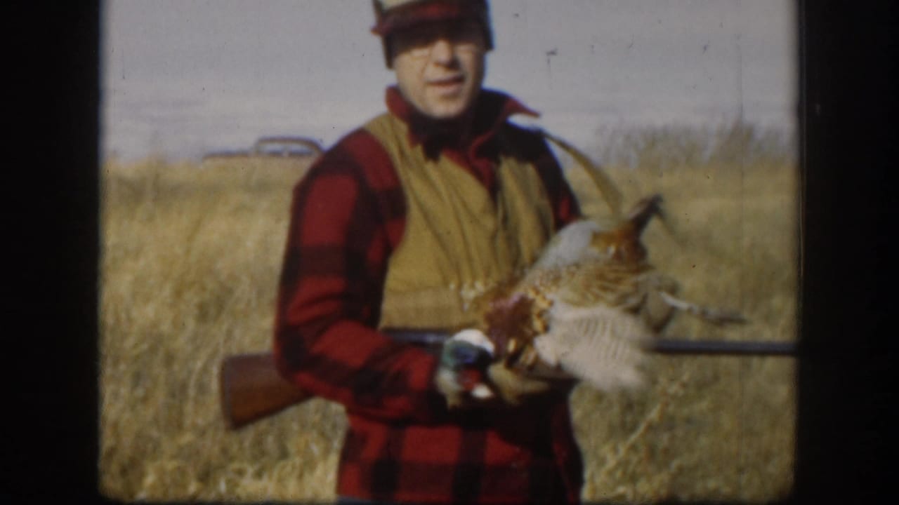 Hunter in red plaid shirt and tan vest raises a pheasant to display its plumage, hunting long gun in hand, dry Kansas prairie and vintage car visible behind him, circa 1958.