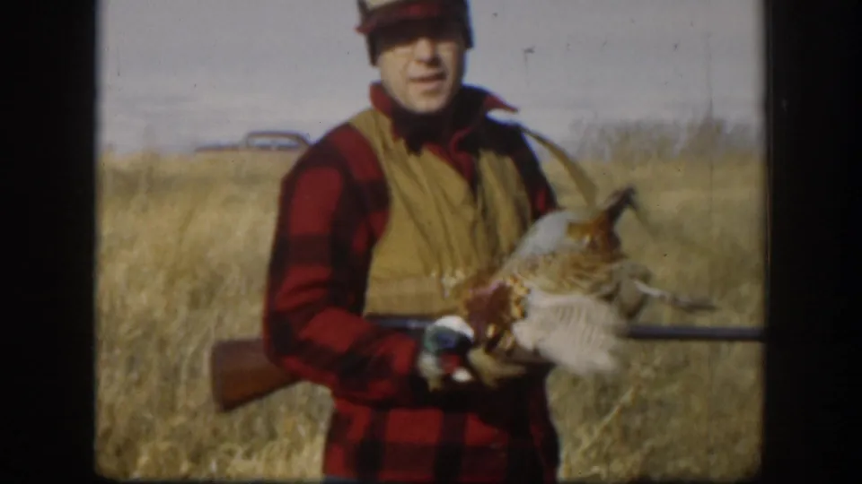 Hunter in red plaid shirt and tan vest raises a pheasant to display its plumage, hunting long gun in hand, dry Kansas prairie and vintage car visible behind him, circa 1958.