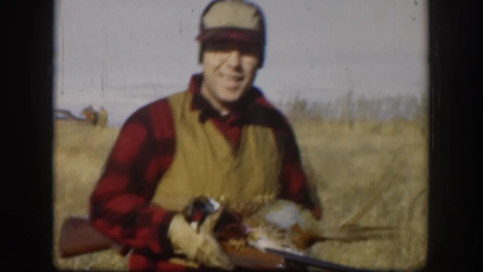 Hunter in red plaid shirt and tan hunting vest smiles toward camera while holding a harvested pheasant, gun resting across his arms, in dry Kansas prairie, circa 1958.