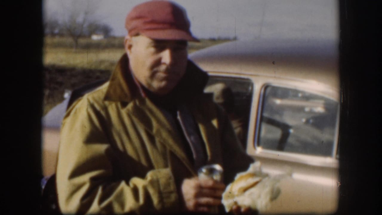 Middle-aged worker in red soft cap and tan work jacket holds a metal beverage can and food item beside a late-1950s light-colored American sedan; flat rural Kansas landscape, circa 1958.