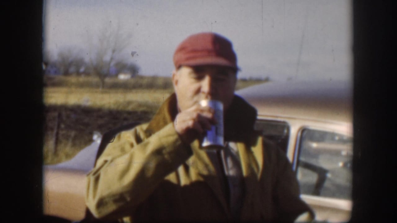 Worker in red soft cap and tan work jacket drinks from a metal can in front of a late-1950s American automobile; flat rural landscape with bare trees visible, Kansas circa 1958.