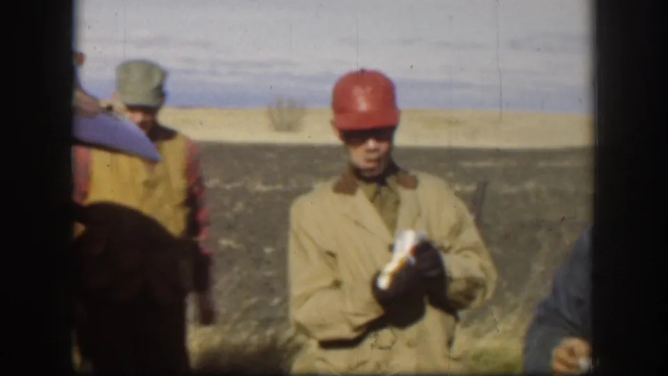 Two outdoor workers during a lunch break — foreground man in red hard hat holds a wrapped food item; second man in grey cap at left; flat open field and partial vehicle visible, Kansas circa 1958.