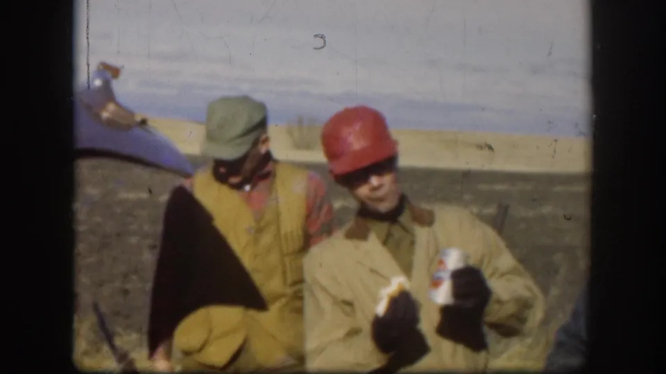 Two male outdoor workers during a lunch break; man in red hard hat holds a food wrapper with possible printed labeling; second man in grey cap at left; flat Kansas-style rural field, circa 1958; film scratch artifact visible.