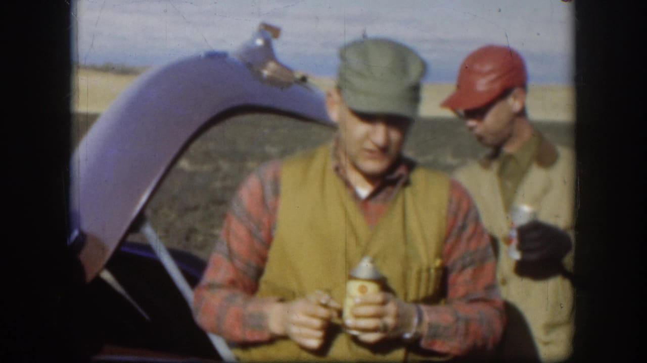 Two adult men in late-1950s workwear stand at an open car trunk in flat terrain; one holds a small can, consistent with Kansas road footage circa 1958.