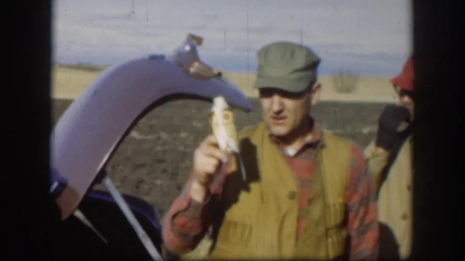 Man in plaid shirt, olive vest and cap holds a white object playfully toward the camera near an open car trunk, with a companion in a red cap behind him, circa 1958 Kansas.