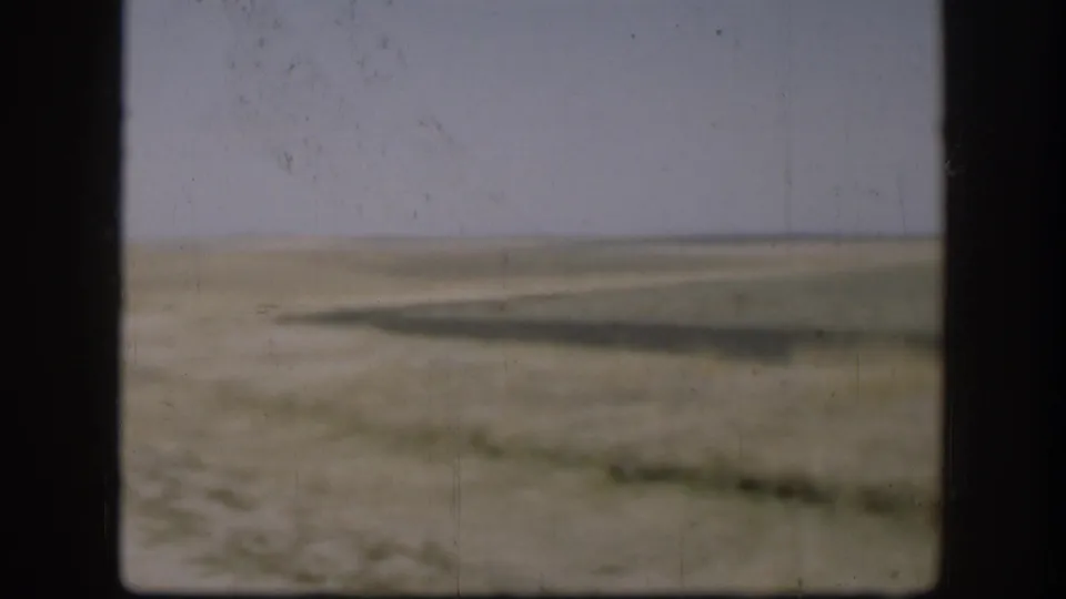 1958 Kansas prairie viewed through a car window: flat treeless grassland, dry brown earth, pale grey-blue sky, and a low flat horizon with no vegetation beyond sparse low grass.