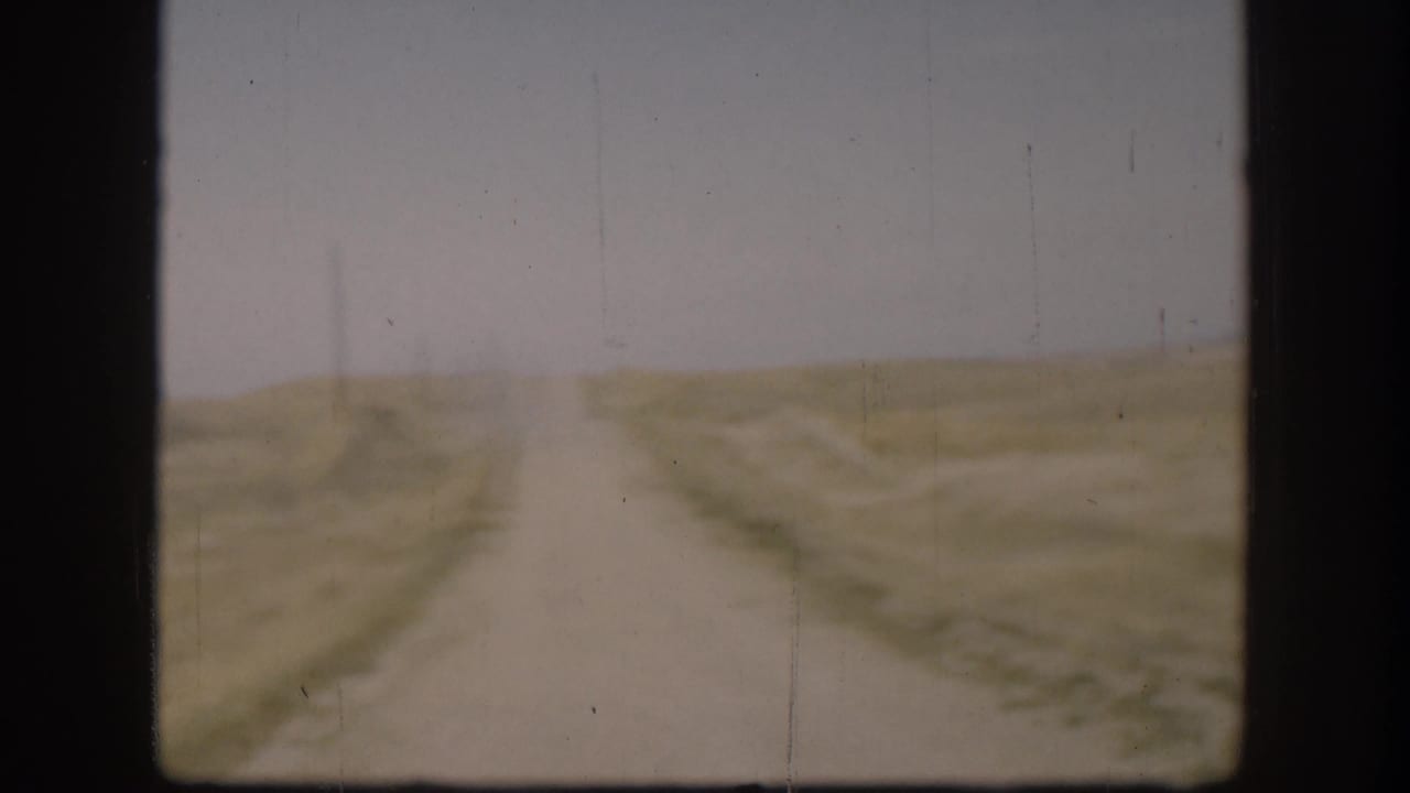 Straight gravel road receding to the flat Kansas horizon, open golden-brown fields on either side, circa 1958 8mm home movie footage.