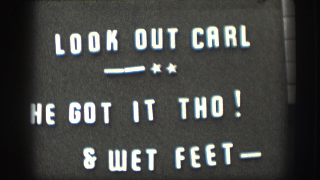 8mm home-movie intertitle card with white hand-lettered text on dark ground: 'LOOK OUT CARL / HE GOT IT THO! / & WET FEET-'; names a family member and narrates a playful outdoor incident.
