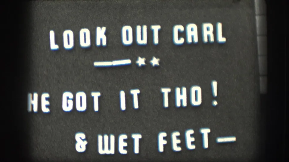 8mm home-movie intertitle card with white hand-lettered text on dark ground: 'LOOK OUT CARL / HE GOT IT THO! / & WET FEET-'; names a family member and narrates a playful outdoor incident.